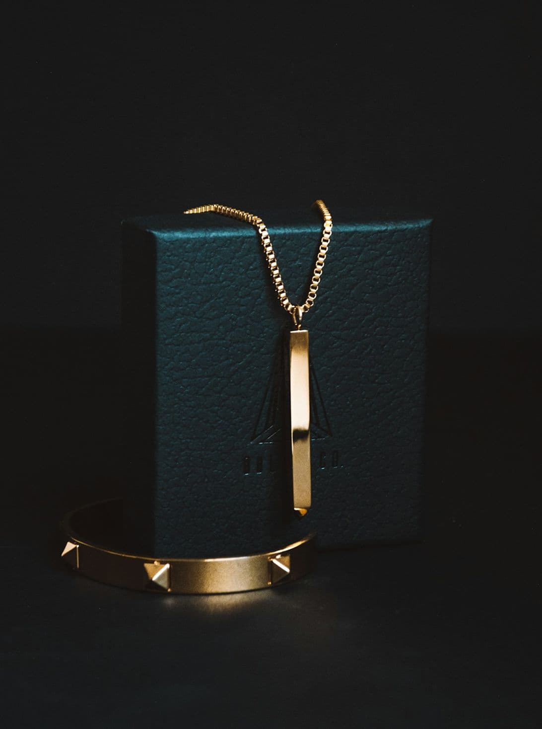 Gold necklace with rectangular pendant and matching spiked bracelet next to a dark teal box.